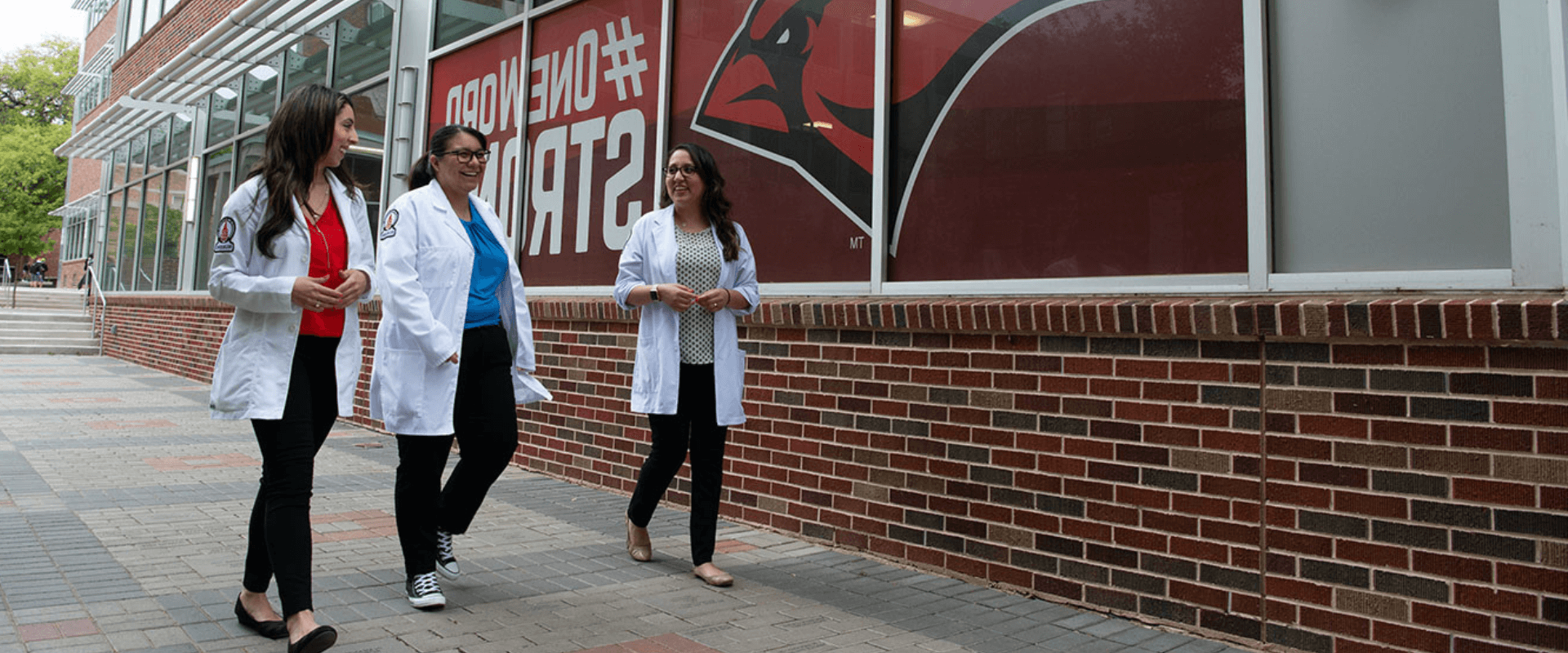 Three MSN Nursing Leadership students walking past a UIW building wearing their Nursing white coats.